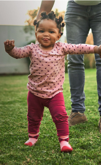 Une petite fille marche dans un jardin, sa main droite est tenue par un adulte.